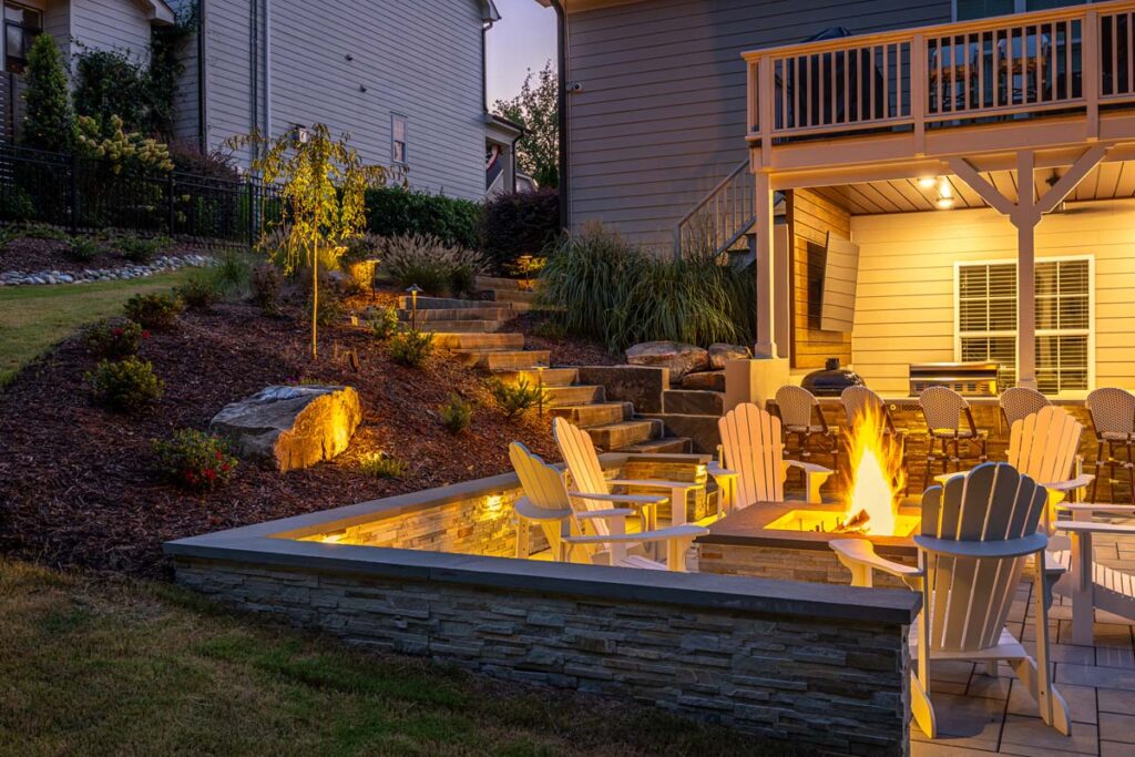 Evening scene showing Roseroot patio surrounded by retaining wall and fire pit. Showcasing patio steps leading to decking and outdoor kitchen the background under composite deck.