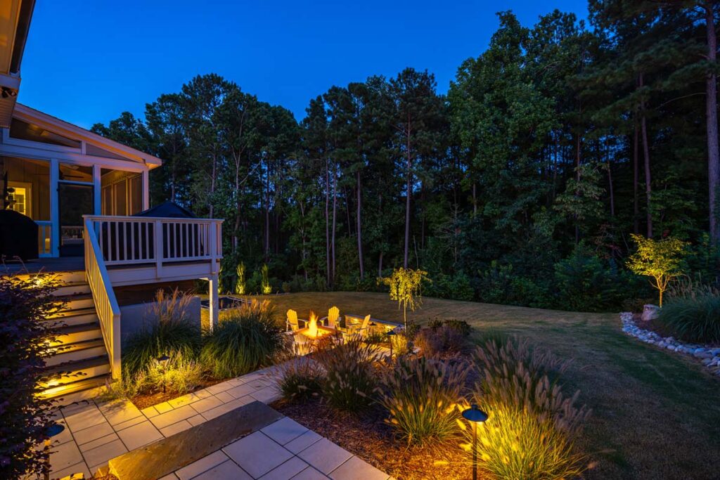 Night scene of Roseroot facing towards forest, showcasing deck leading to fire pit, featuring low-voltage lighting.