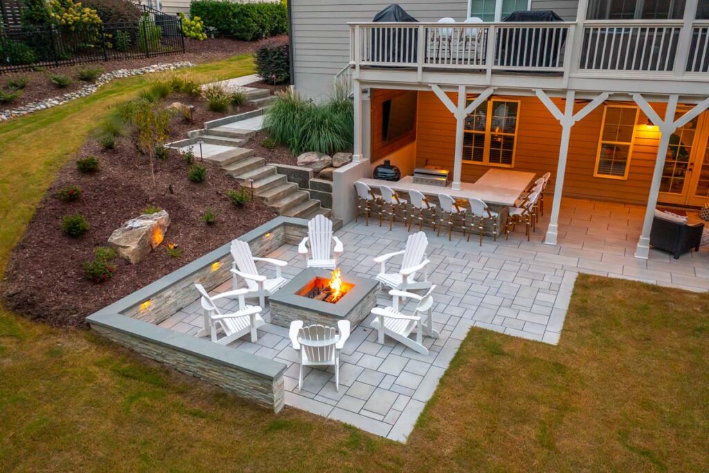 Top angled view facing Roseroot deck with pathway leading to patio with fire place, featuring products from TimberTech and TechoBloc. Showing more outdoor kitchen underneath deck.