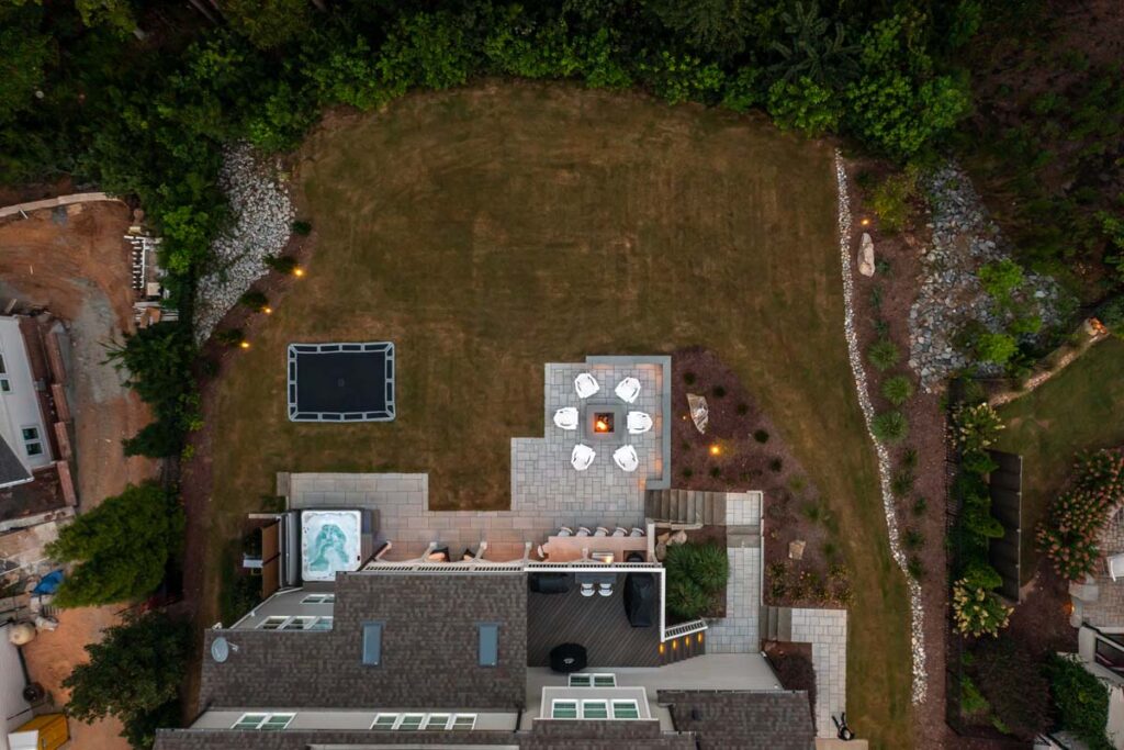 Zoomed out aerial view of Roseroot showing patio with fireplace, deck, walkways and low-voltage lighting. Showing more of neighboring properties.