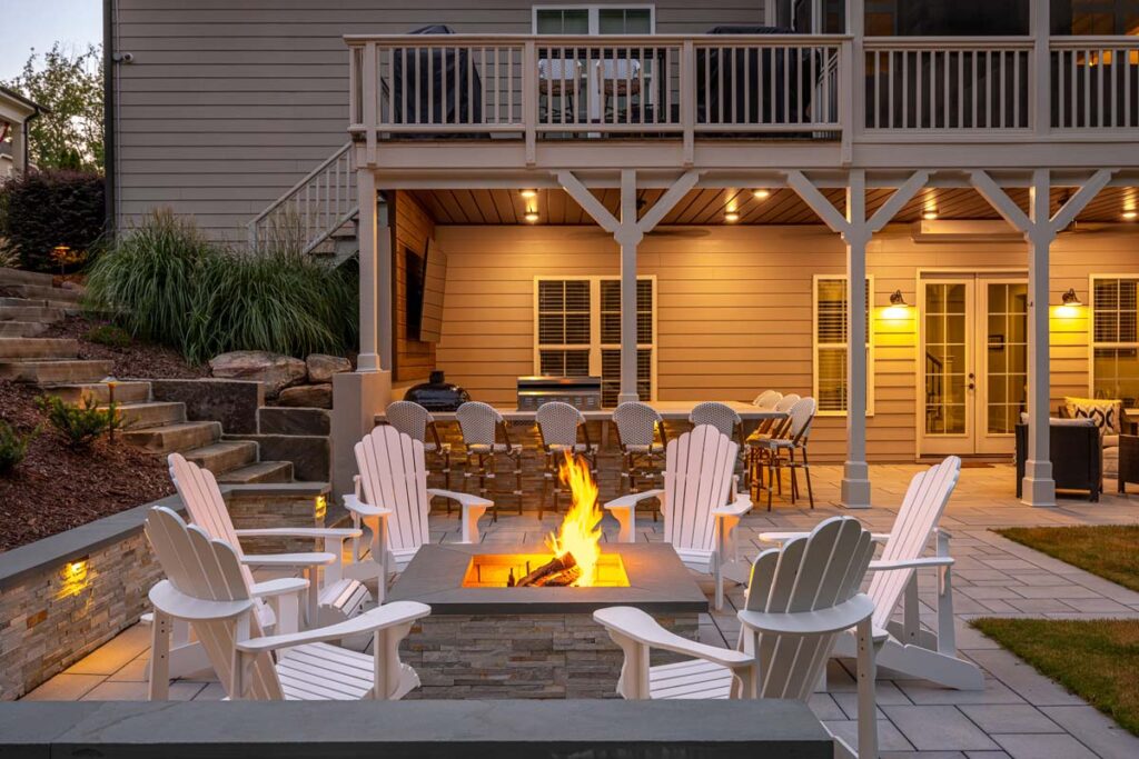 A direct front-facing evening scene of Roseroot, showing patio fire pit in the foreground and outdoor kitchen in the background.