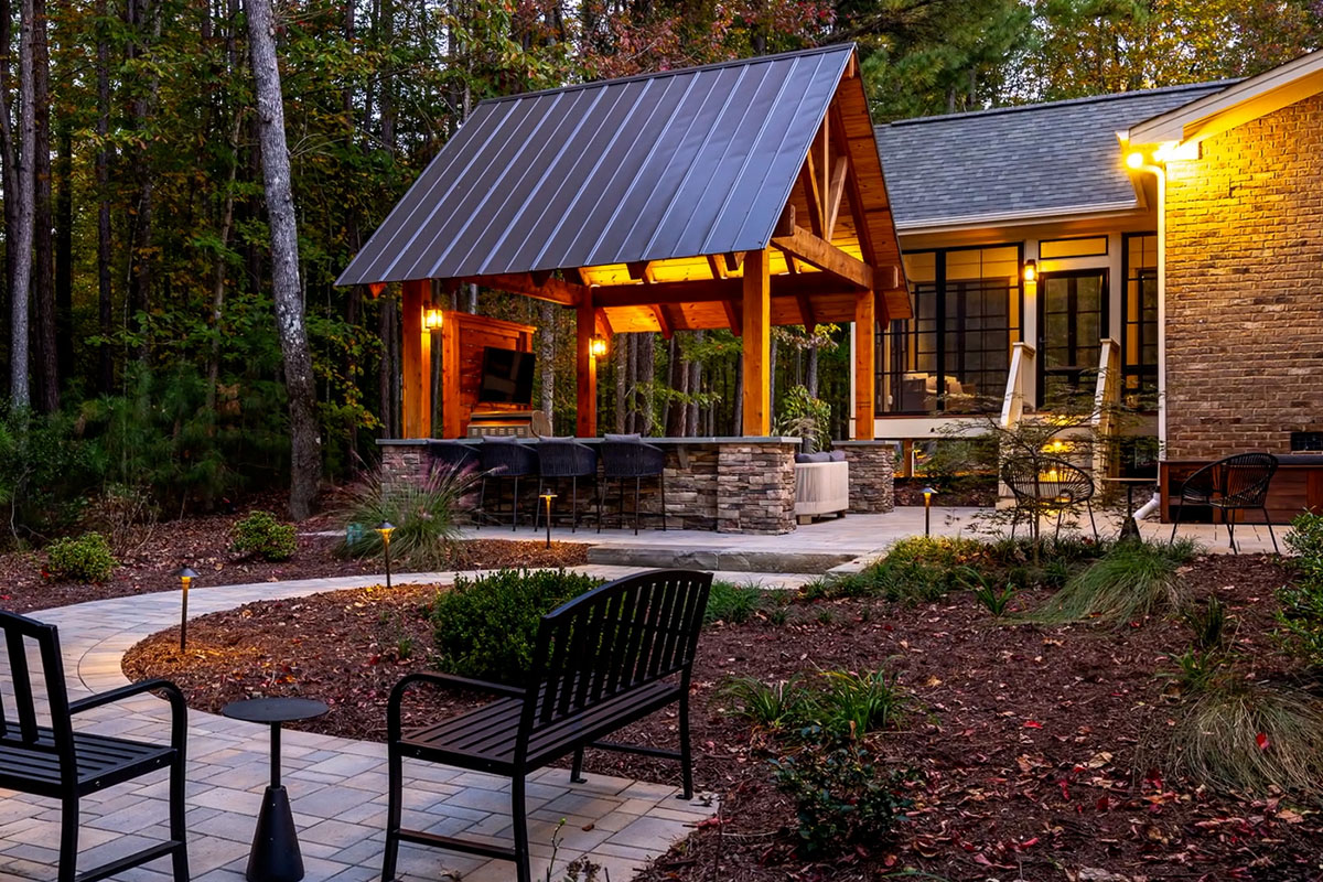 After: Side view of finished Colvard Park Pavilion and home extend with an screened deck. Pavilion is fully finished with a tv and counter top area with stools. Walkway is finished and covered with garden shrubs. Close to the camera shows two metal benches.