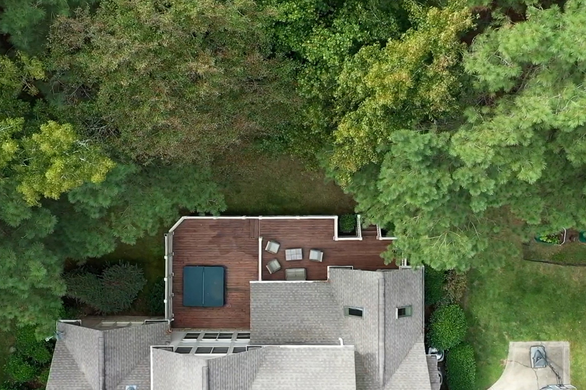 Before: Top view of Colvard Park home with a wooden large wooden deck that has a seating area and jacuzzi. Surrounded by shrubs and trees.