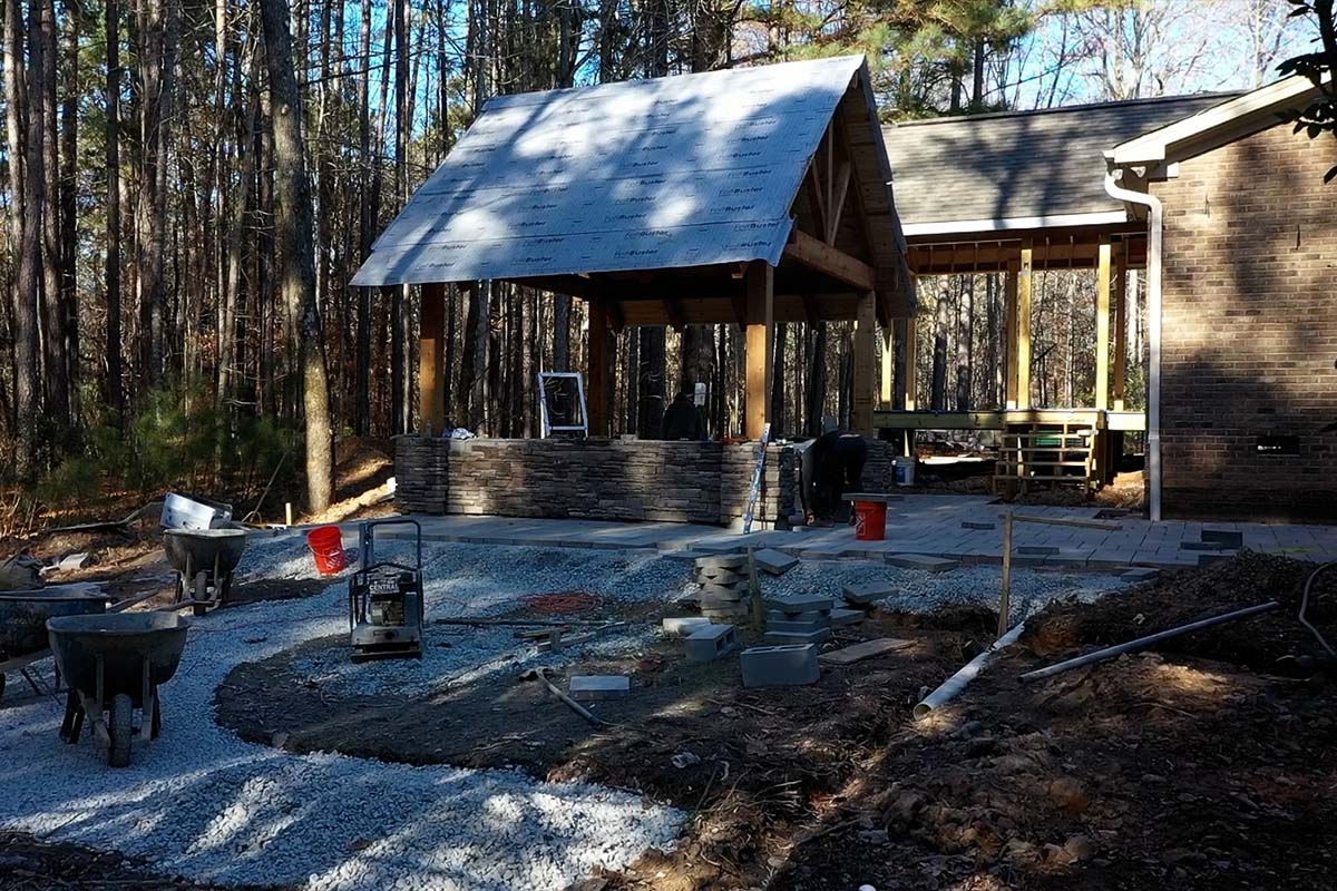 Before: Side view of in-progress Colvard Park with in progress pavilion, with missing shingles and fireplace. Walkway is unfinished showing gravel used to grade the ground before applying the pavers. Ground is open to reveal drainage system under walkway.