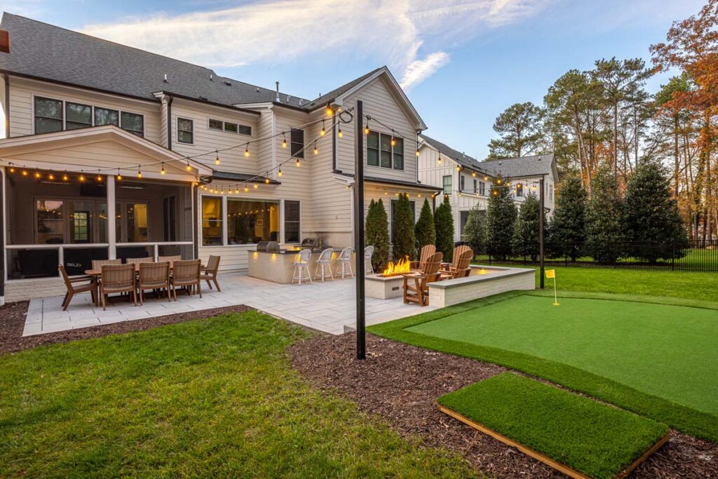 Afternoon wide shot of Skybrook Oak patio with patio table on the left, outdoor kitchen in the center, fire pit on the right, and golf area in the foreground.